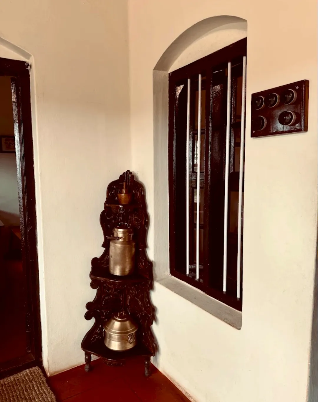 An arched window with dark wooden shutters, brass vessels stacked on a carved stand against a red-oxide floor.